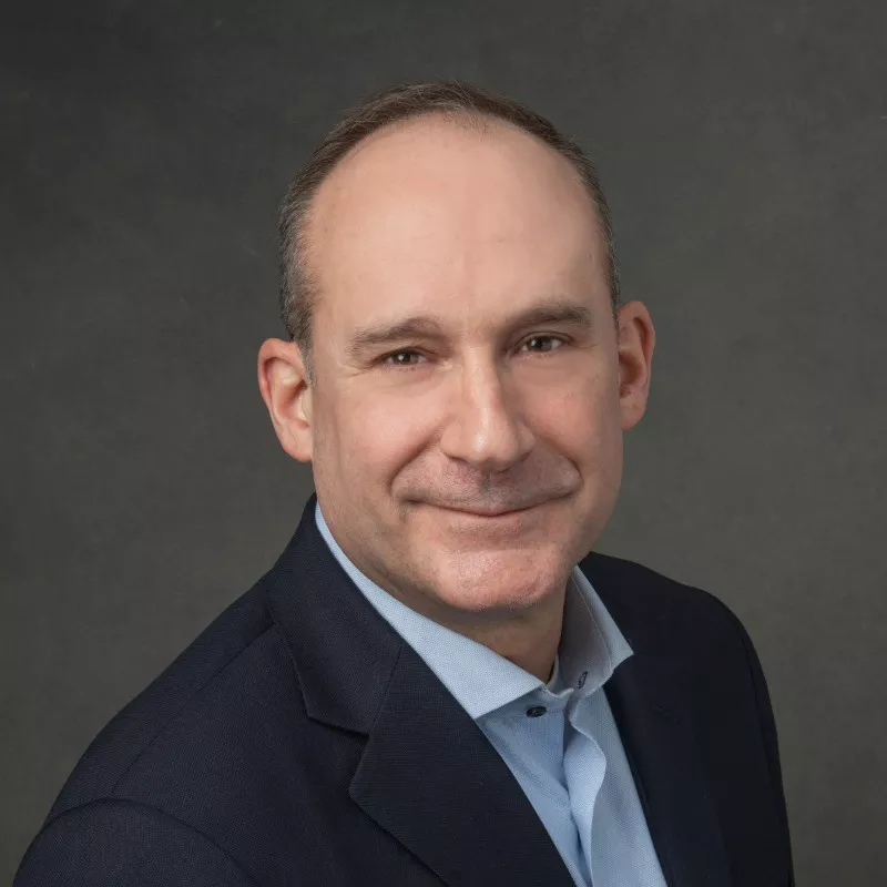 A professional man is dressed in a dark suit jacket and light blue collared shirt, set against a neutral gray background. The image conveys a formal and polished appearance, suitable for corporate or professional contexts. The overall tone is neutral and sophisticated.